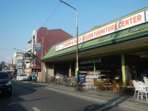 Eine belebte Stadtstraße mit fahrenden Fahrzeugen, parkenden Fahrrädern, Gebäuden mit Schildern, Laternenmasten und Stromleitungen, mit einem Möbelgeschäft im Vordergrund und dem Himmel im Hintergrund.