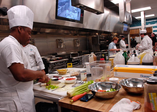 Eine Gruppe von Köchen in einer Küche, die auf einem Schneidbrett Essen zubereiten, mit einem Koch im Vordergrund in einem weißen Kleid und einer Mütze, der ein Messer hält und von Utensilien, Schüsseln, Gemüse, einem Handy, Flaschen und anderen Gegenständen umgeben ist, mit einem Fernseher und Deckenlampen im Hintergrund.