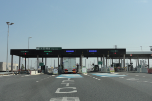 Eine Mautstation am Straßenrand mit einem vor ihr geparkten Lkw, weiße und blaue Straßenmarkierungen, Laternenpfähle, Schilder und andere Gegenstände auf beiden Seiten, mit Gebäuden, Bäumen und einem klaren blauen Himmel im Hintergrund.