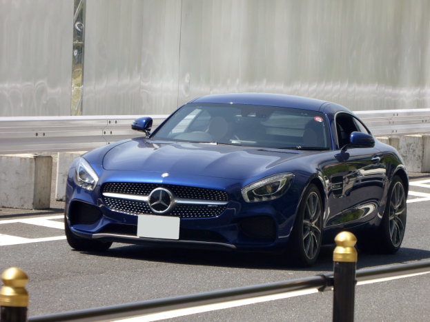 Ein blaues Mercedes-Benz AMG GT Coupé mit schlanker, aerodynamischer Karosserie, eckigen Scheinwerfern, großem Kühlergrill, glänzenden Rädern und luxuriösem Innenraum, das eine Straße entlangfährt, mit einer Wand im Hintergrund.