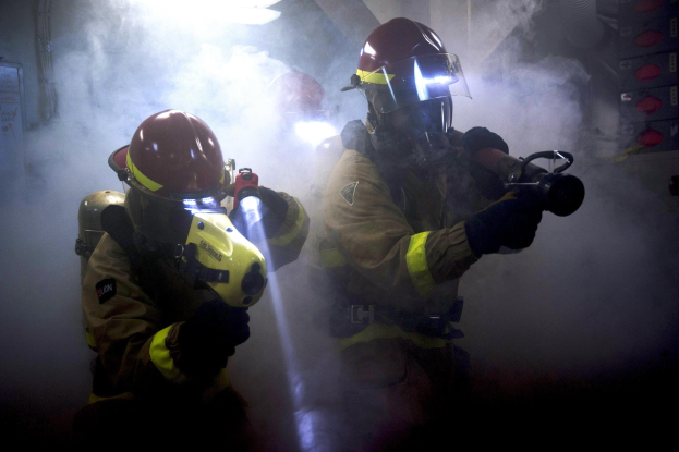 Zwei Feuerwehrleute in Schutzausrütstüßen spritzen Wasser auf eine rauchgefüllte Wand, mit einer Platte und verschiedenen Gegenständen in der Nähe.