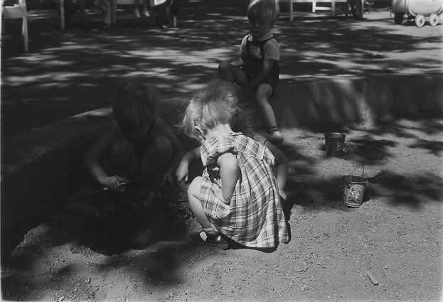 Ein Schwarz-Weiß-Foto von drei Kindern, die im Sand spielen, mit einem Jungen und einem Mödchen, die neben einem Eimer sitzen, und einem Wagen rechts daneben.