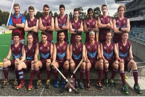 Eine Gruppe von Männern, die auf einem Feld mit Hockeystöcken feiern, umgeben von Bäumen und einem blauen Himmel, und ein Stadion mit Zuschauern auf der rechten Seite.