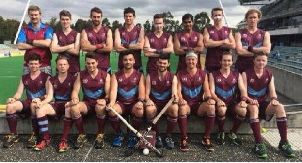 Eine Gruppe von Männern, die auf einem Feld mit Hockeystöcken feiern, umgeben von Bäumen und einem blauen Himmel, und ein Stadion mit Zuschauern auf der rechten Seite.