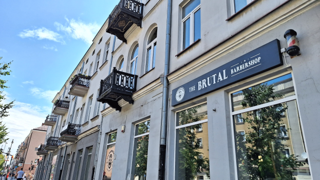 Brutalistischer Friseurladen in Berlin mit Betongebäuden, Fenstern, Geländern, Balkonen und Namensschildern, umgeben von Bäumen, Straßenlaternen und Fußgängern unter einem bewölkten Himmel.