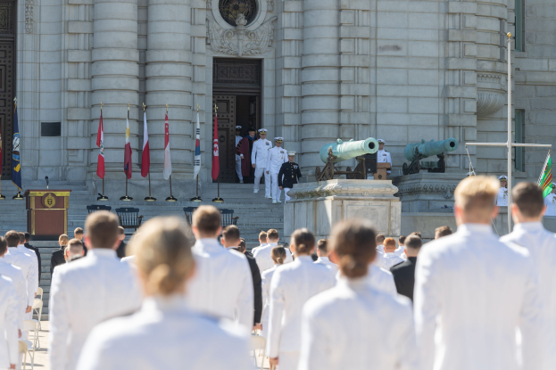 Gruppe von Menschen in weißen Marineuniformen, die vor einem Gebäude mit Säulen und einer Tür stehen, mit Fahnen, einem Podium und Kanonen im Hintergrund.