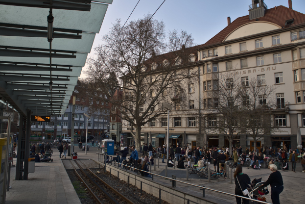 Eine belebte Straßenkreuzung mit einem Bahnhof auf der linken Seite, umgeben von Gebäuden, Bäumen und Laternen, wo Menschen gehen, auf Bänken sitzen oder in der Nähe des Bahnhofs stehen.