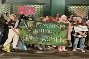 Eine Gruppe von Menschen mit einem Banner, auf dem "Kein Feminismus ohne Trans-Frauen" steht und verschiedenen Schildern in einem öffentlichen Raum.