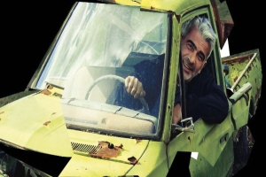 Ein Mann, der in einem gelben, rostigen und verbeulten Auto auf dem Fahrersitz sitzt und lächelt.