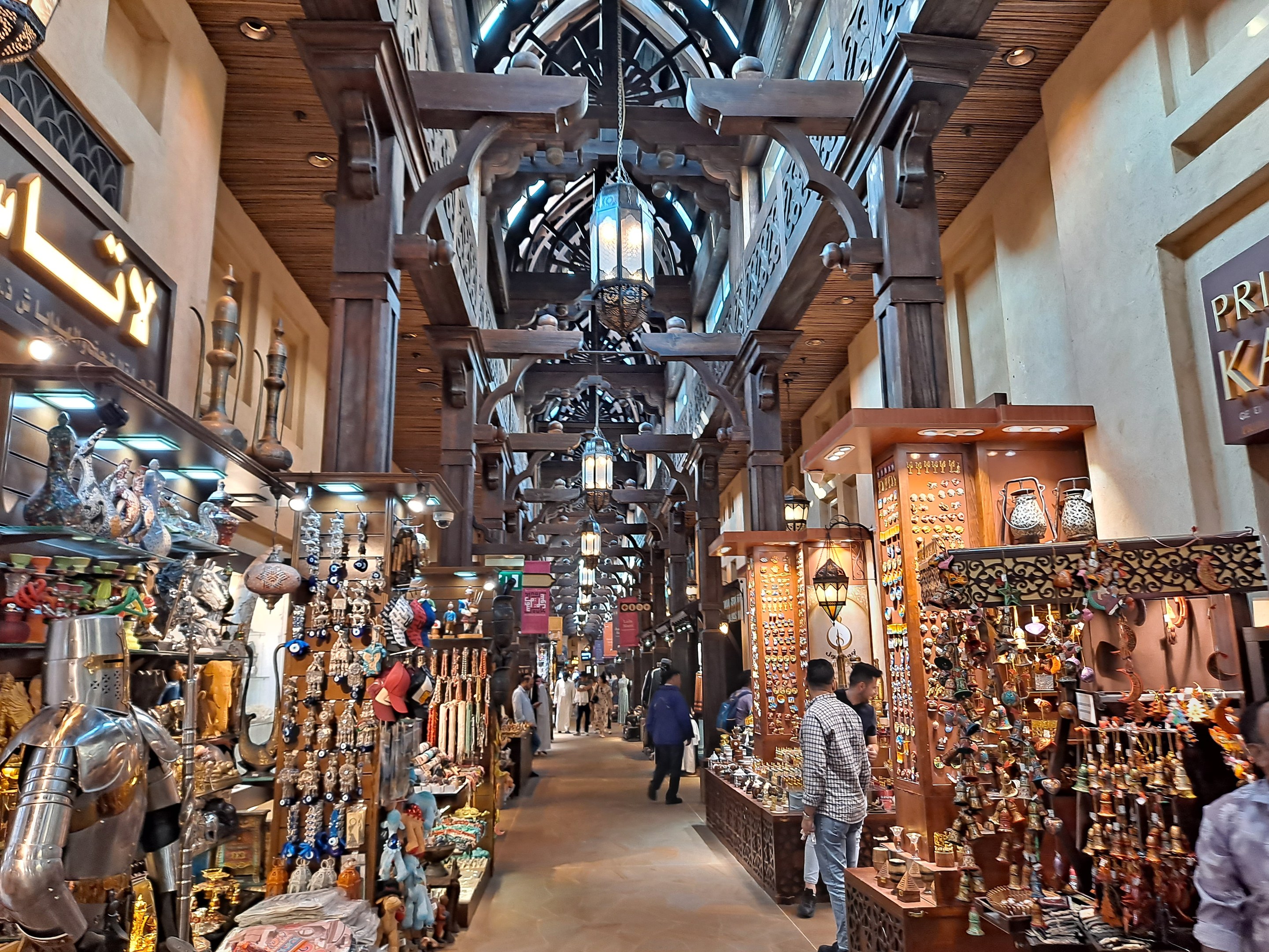 Ein geschäftiges Dubai-Basar mit Menschen, die zwischen Ständern mit Waren hindurchgehen, Wänden mit Texttafeln und einer Decke mit warmem Licht.