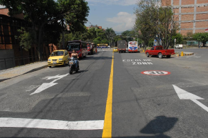 Eine Straßenszene mit Gebäuden, Bäumen, fahrenden Fahrzeugen, Verkehrsschildern, Ampeln und Warnmarkierungen unter einem blauen bewölkten Himmel.