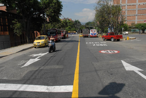 Eine Straßenszene mit Gebäuden, Bäumen, fahrenden Fahrzeugen, Verkehrsschildern, Ampeln und Warnmarkierungen unter einem blauen bewölkten Himmel.