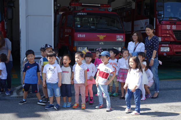 Eine Gruppe von Kindern vor mehreren Feuerwehrautos an einer Feuerwache, einige tragen Mützen.
