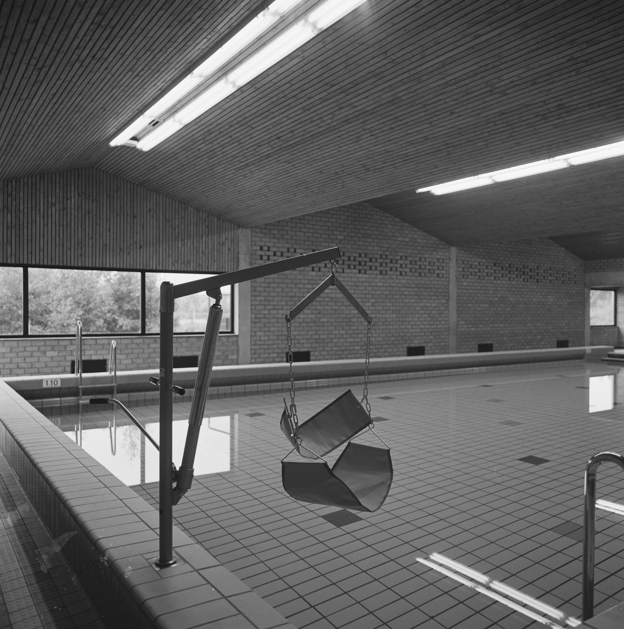 Schwarz-weiß-Foto eines Hallenbades mit einer von der Decke hängenden Schaukel, umgeben von einem gefliesten Boden, Metallstangen und Glasfenstern, die auf Bäume hinausgehen, mit Deckenlampen, die eine gemütliche Atmosphäre schaffen.