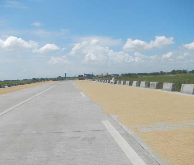 Eine Straße mit Betonleitwänden auf beiden Seiten, Fahrzeuge, die darauf fahren, und Bäume und Wolken im Hintergrund.