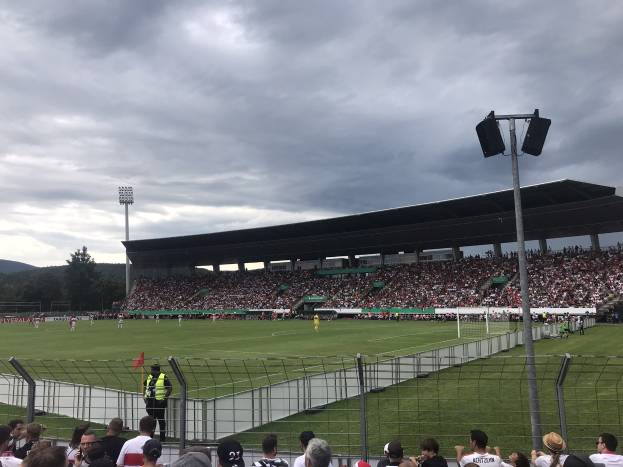 Große Zuschauermenge bei einem Fußballspiel in einem eingezäunten Stadion unter Flutlichtmasten, mit Bäumen und Hügeln im Hintergrund unter einem bewölkten Himmel.