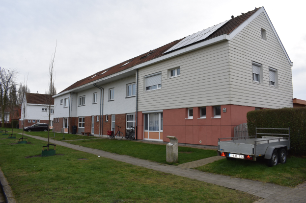 Ein Lastwagen, der vor einer Reihe von Häusern mit Solarpaneelen auf dem Dach geparkt ist, Fahrräder in der Nähe und ein grasbewachsener Weg, der zu den Häusern führt, mit Bäumen und einem bewölkten Himmel im Hintergrund.