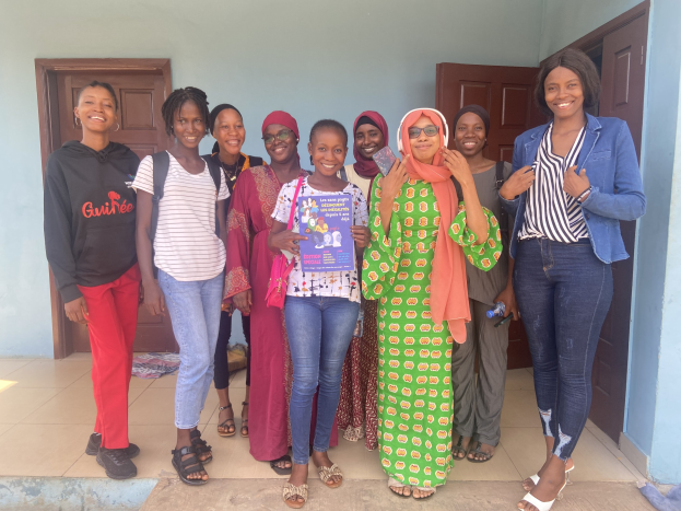 Eine Gruppe von Frauen vor einem Schulgebäude in Nigeria, die strahlend Bücher halten und ein Tuch auf dem Boden drapiert haben, mit Türen im Hintergrund.