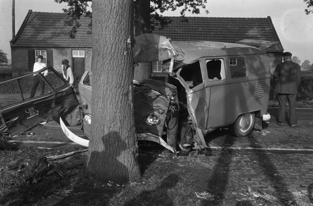 Schwarz-weißes Foto eines zerbeulten Autos am Straßenrand mit einem Baum im Vordergrund und einigen Menschen in der Nähe, Häusern und Bäumen im Hintergrund.