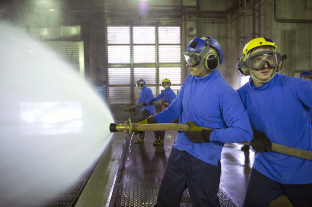 Gruppe von Männern in blauen Hemden und gelben Helmen bei der Arbeit an einer Maschine in einer Fabrik mit Rohren, Fenstern und Lampen.