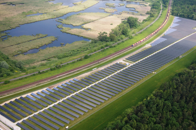 Luftaufnahme einer großen Solar-Farm auf einem Feld mit Bäumen, Gras, Pflanzen und Wasser, mit Solar-Panelen und einem Zug auf einer Bahnstrecke.