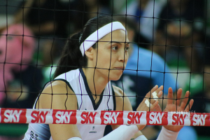 Eine weibliche Volleyballspielerin in Uniform ist auf dem Feld im Einsatz, mit einem Netz im Vordergrund und einem unscharfen Hintergrund, mit einem entschlossenen Gesichtsausdruck.