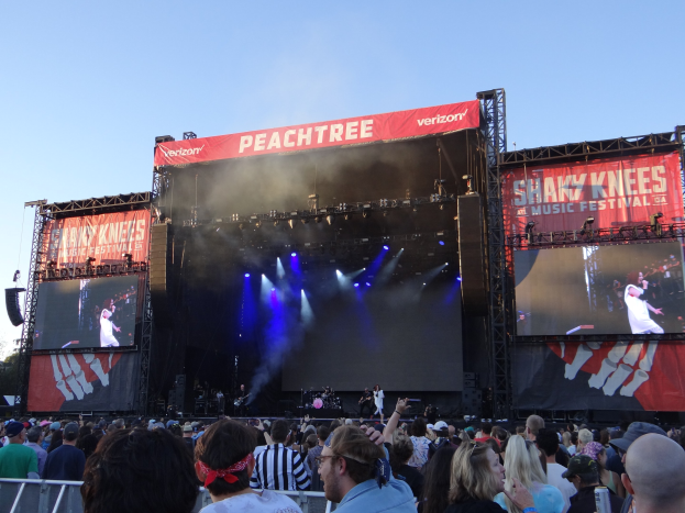 Eine große Menschenmenge steht vor einer hell erleuchteten Bühne auf dem Shaky Knees Music Festival, wo Musiker hinter einem Geländer unter einem klaren blauen Himmel spielen.