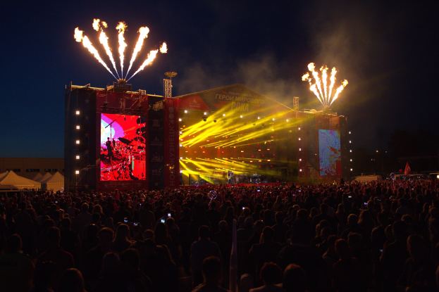 Eine große Menschenmenge auf einem Musikfestival mit einer Bühne, auf der Musiker unter hellen Lichtern spielen, umgeben von Bannern, Zelten links daneben und einem dunklen Himmel mit Feuerwerk dahinter.