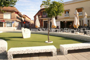 Minigolfanlage in einem Stadtplatz umgeben von Sitzgelegenheiten, Sonnenschirmen, Bäumen und Gebäuden unter einem bewölkten Himmel.