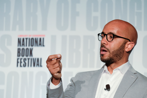 Ein Mann in Anzug und Brille spricht an einem Podium mit einem "National Book Festival"-Schild dahinter und trägt ein Mikrofon am Hemd.