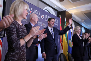 Eine Gruppe von Menschen klatscht feierlich vor Flaggen, mit einem Tisch voller Gegenstände rechts daneben, Stühlen, einer Fahne und einer Wand im Hintergrund sowie Lampen und einem Ventilator auf dem Dach.