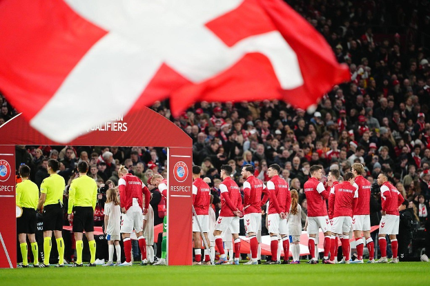 Gruppe von Menschen auf einem Fußballfeld mit einer roten und weißen Flagge im Vordergrund, einem Bogen mit der Aufschrift "Bayern München vs. Wetten und Vorschau für Bayern München" im Hintergrund und einer großen Menschenmenge im Stadion.