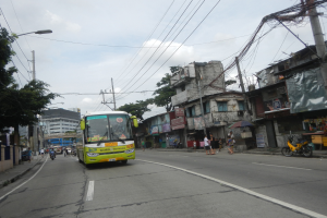 Ein grüner Bus fährt auf einer Straße mit hohen Gebäuden, Strommasten, Laternen, Bäumen und Menschen, die zu Fuß gehen oder Fahrrad fahren, unter einem bewölkten Himmel.