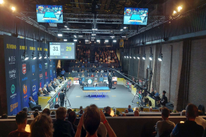 Eine große Menschenmenge sitzt vor einer Bühne mit einem Tischtennis-Tisch in der Mitte, Stühlen auf der linken Seite, einer Wand auf der rechten Seite und Deckenlampen und Fernsehern im Hintergrund bei der 2019 Jumbo-Weltmeisterschaft.