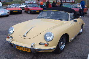 Ein gelber Porsche 356 Speedster auf einem Parkplatz mit Menschen in der Nähe, vor einer Kulisse aus einem Zaun, Bannern, Bäumen, Gebäuden und einem klaren Himmel.