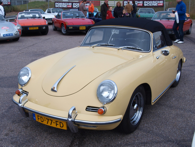 Ein gelber Porsche 356 Speedster auf einem Parkplatz mit Menschen in der Nähe, vor einer Kulisse aus einem Zaun, Bannern, Bäumen, Gebäuden und einem klaren Himmel.
