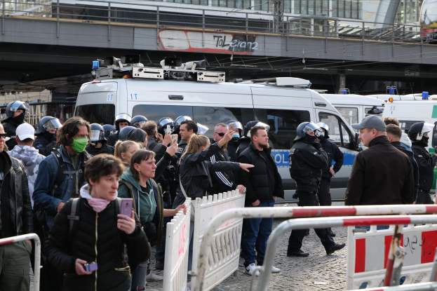 Eine Gruppe von Menschen steht vor Polizeifahrzeugen in Berlin, einige tragen Helme und halten Telefone, mit Barrieren im Vordergrund und einer Brücke mit Gebäuden im Hintergrund.