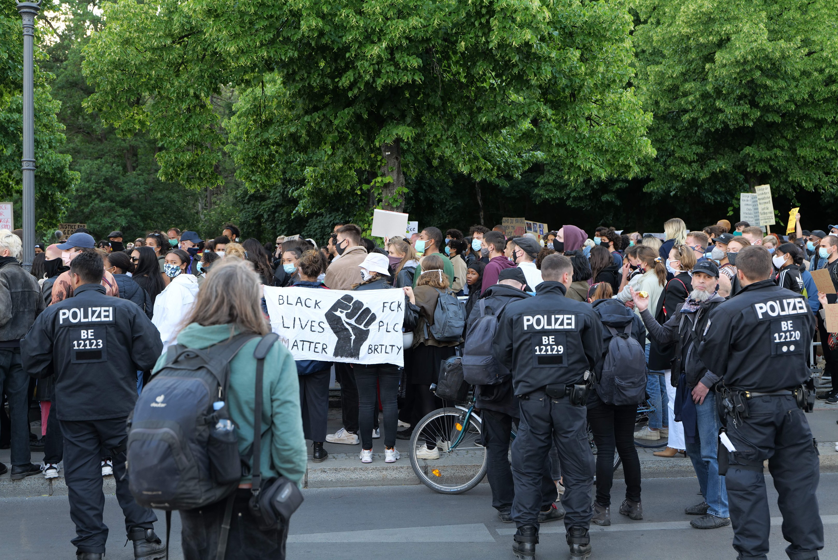 Eine große Gruppe von Menschen steht an der Straße, einige halten Schilder und andere tragen Mützen und Taschen, vorne ein Fahrrad und im Hintergrund Bäume und ein Pfahl.
