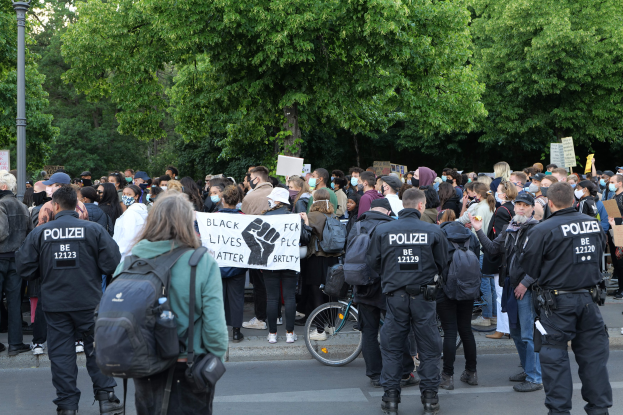 Eine große Gruppe von Menschen steht an der Straße, einige halten Schilder und andere tragen Mützen und Taschen, vorne ein Fahrrad und im Hintergrund Bäume und ein Pfahl.