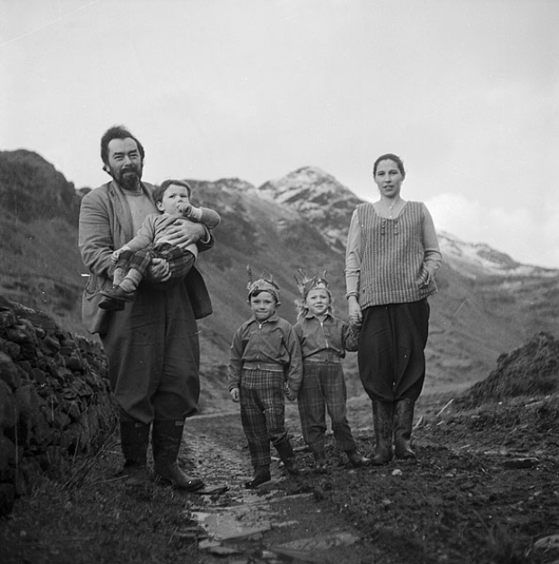 Eine Familie von vier Personen steht auf einem Schotterweg in den Bergen, wobei der Mann ein Baby hält, die Frau und zwei Kinder neben ihm stehen, vor dem Hintergrund von Hügeln und einem bewölkten Himmel dargestellt in Schwarz-Weiß.
