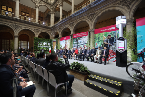 Eine Gruppe von Menschen sitzt in Stühlen und schaut auf einen Mann auf einer Bühne, der ein Mikrofon hält, rechts davon stehen verschiedene Fahrräder und im Hintergrund sind Werbetafeln, Pflanzen, Säulen, Geländer, Lampen, Fenster und eine Wand zu sehen.