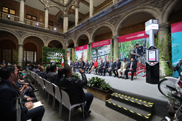 Eine Gruppe von Menschen sitzt in Stühlen und schaut auf einen Mann auf einer Bühne, der ein Mikrofon hält, rechts davon stehen verschiedene Fahrräder und im Hintergrund sind Werbetafeln, Pflanzen, Säulen, Geländer, Lampen, Fenster und eine Wand zu sehen.