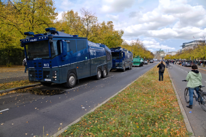 Eine Gruppe von Polizeifahrzeugen, die auf der Seite einer Straße geparkt sind, mit einer Person, die auf einem Fahrrad auf der rechten Seite fährt, umgeben von vielen Menschen, einige halten Fahrräder, mit Gras, trockenen Blättern, Bäumen, Gebäuden und einem bewölkten Himmel im Hintergrund.