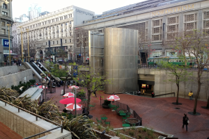 Eine belebte Stadtstraße mit Fußgängern, Regenschirmen, Außensitzgelegenheiten, Grünflächen und einem großen Einkaufszentrum im Hintergrund unter einem sichtbaren Himmel.