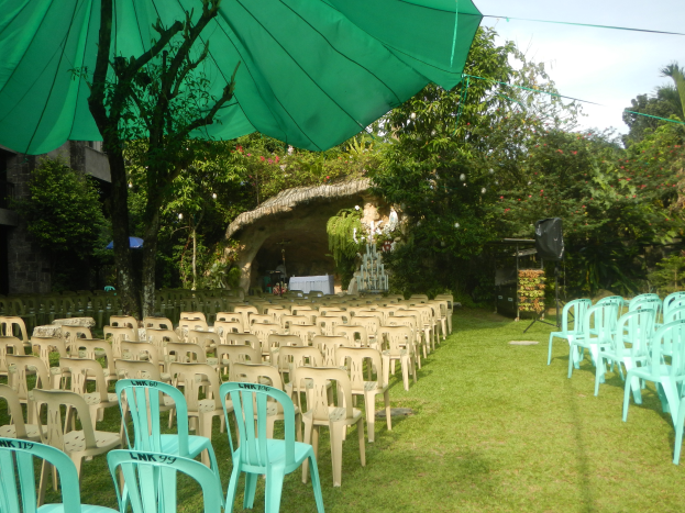 Eine Hochzeitszeremonie im Garten mit grünen Stühlen und Schirmen, umgeben von üppiger Vegetation, mit einem Gebäude und einem Schuppen im Hintergrund und dem Himmel oben sichtbar.