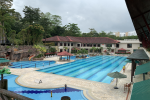 Großes Schwimmbad im Ritz-Carlton, Kota Kinabalu, mit Menschen, Sonnenschirmen, Stühlen, Tischen, Pflanzen, Bäumen, Felsen, Gebäuden und Himmel.