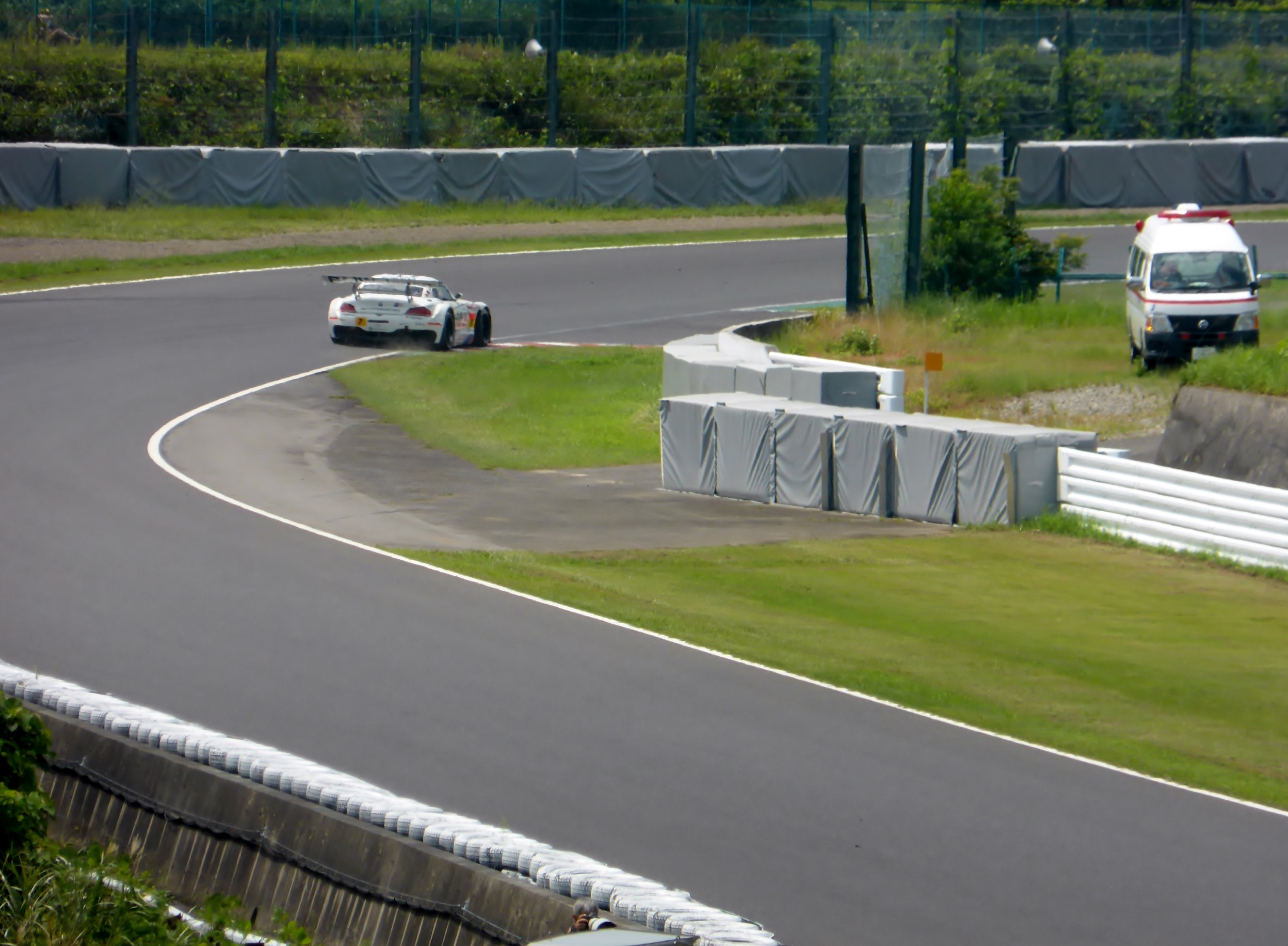 Ein Rennwagen auf einer Rennstrecke mit einem Polizeiwagen im Hintergrund, umgeben von Gras, Pflanzen, Bäumen, Zäunung und einem Fahrzeug auf der rechten Seite des Bildes.