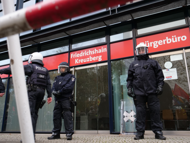 Eine Gruppe von Polizisten in Helmen und Handschuhen steht vor einem Glasgebäude mit einer Stange links und Schildern im Hintergrund, die 'Friedrichshain Kreuzberg' und Papiere an den Glaswänden angeklebt zeigen.
