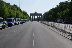Lange Reihe von Polizeiwagen, die auf der Seite einer Straße vor dem Brandenburger Tor in Berlin, Deutschland, geparkt sind, mit Menschen, die Fahrräder fahren und auf der Straße stehen, Barrieren, Bäumen, einem Bogen mit Statuen im Hintergrund und sichtbarem Himmel.