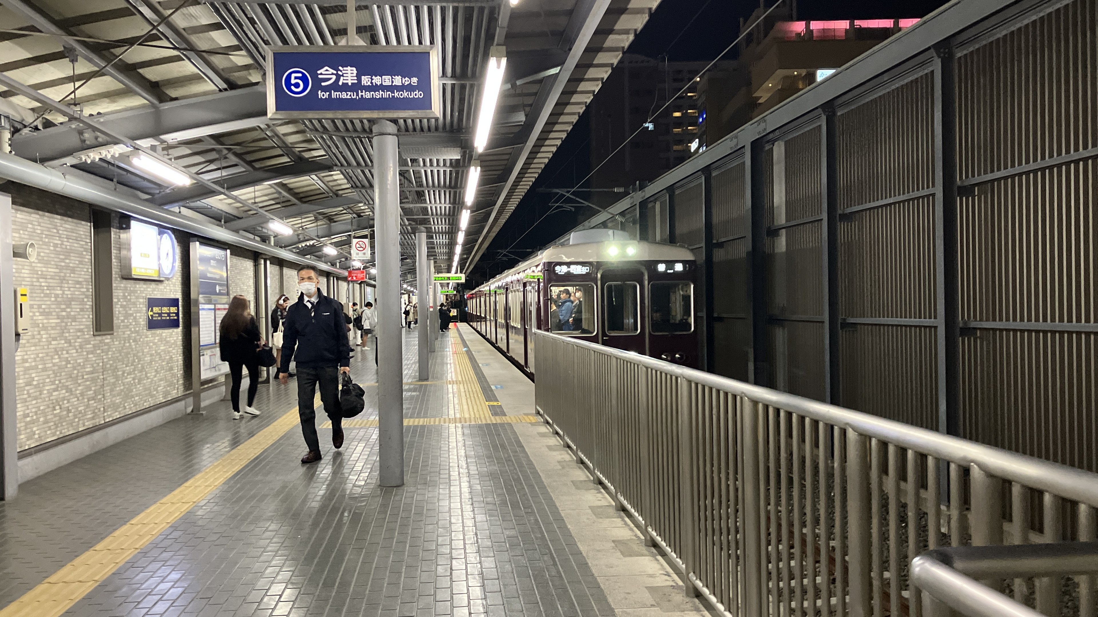 Ein Zug fährt auf einem Bahnsteig ein, mit Menschen in der Nähe, einem Geländer auf der rechten Seite, Gebäuden im Hintergrund, Bahnsteiglütung und einer Anzeigetafel.